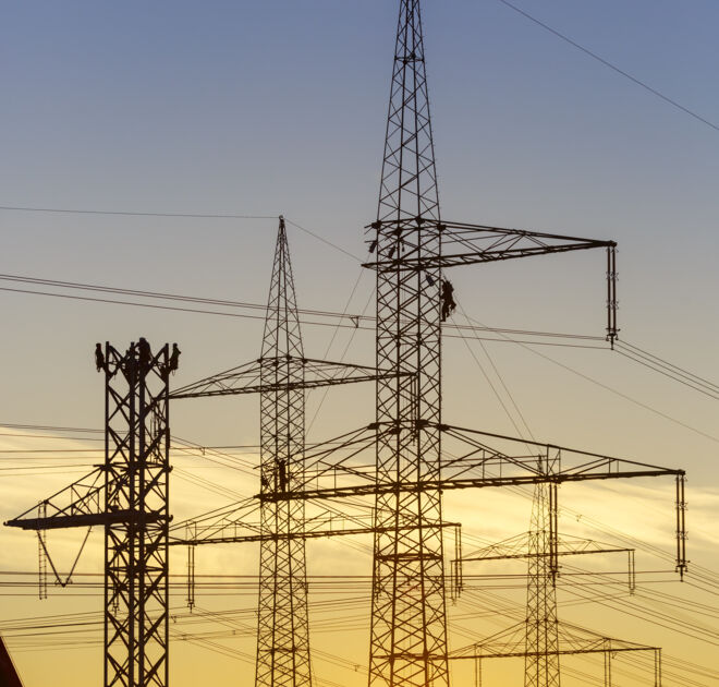 Stromleitungen und Freileitungsmasten im Sonnenuntergang mit Monteuren bei Arbeiten an der Leitung.